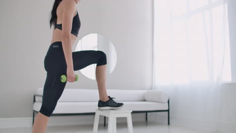 A-young-brunette-woman-performs-concentrated-pacing-on-a-chair-in-a-bright-interior-apartment-with-dumbbells-in-her-hands.